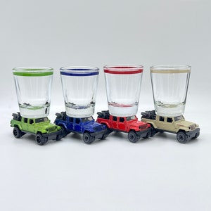 May include: Four shot glasses, each with a different colored rim, are stacked on top of toy Jeep Wrangler vehicles. The glasses are green, blue, red, and clear. The Jeeps are all black and have a silver grill.