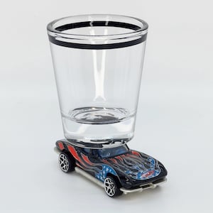 May include: A black and red toy car with a blue and white American flag design is being held up by a clear shot glass with a black band around the rim.