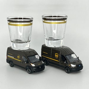 Könnte beinhalten: Zwei klare Schnapsgläser mit schwarzen und goldenen Bändern stehen auf braunen UPS-Lieferwagen-Spielzeugmodellen. Die Transporter haben das UPS-Logo und den Text. Die Schnapsgläser sind leer und haben ein schlichtes Design.