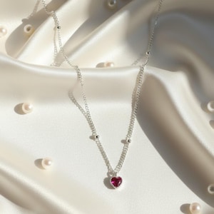 May include: A silver necklace featuring a heart-shaped, deep red pendant. The pendant is framed by a silver border. The chain has small silver beads. The necklace is presented on a white satin background, with scattered pearls.