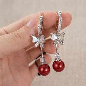 May include: A pair of silver-toned drop earrings featuring a butterfly motif with iridescent white inlay, accented by clear pavé crystals on the hoop and a single round crystal above a smooth, deep red spherical bead at the base.