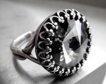May include: A silver ring with a large, faceted, gray gemstone. The gemstone is set in a decorative, filigree-style bezel. The ring is on a silver surface, and the light reflects off the facets of the stone.