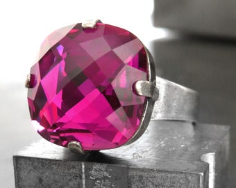 May include: A silver ring with a large, faceted, fuchsia-colored gemstone. The ring is sitting on a gray, textured surface.