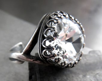May include: A silver ring with a large, clear, faceted gemstone. The ring has an ornate, filigree setting around the stone and a detailed band. The gemstone is square-shaped and reflects light, creating a sparkling effect. The ring is set against a gray background.