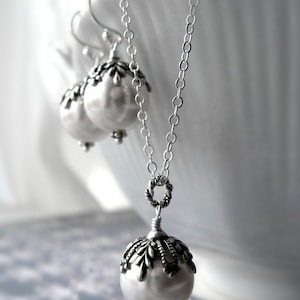 May include: A silver necklace with a large pearl pendant and a silver chain. The pendant has a detailed design around the pearl. The necklace is displayed on a white surface. Earrings & necklace sold separately.