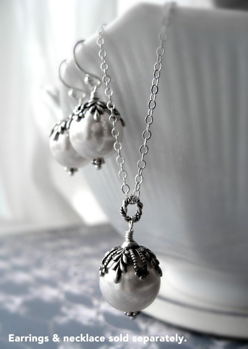 May include: A silver necklace with a large pearl pendant and a silver chain. The pendant has a detailed design around the pearl. The necklace is displayed on a white surface. Earrings & necklace sold separately.
