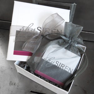 May include: A white gift box with a gray and pink ribbon. The box is open and contains a small gray bag with the text "shySIREN" printed on it.