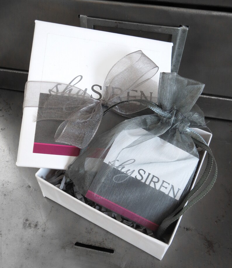 May include: A white gift box with a gray and pink ribbon. The box is open and contains a small gray bag with the text "shySIREN" printed on it.