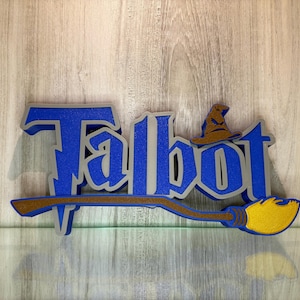 May include: A blue and silver 3D sign with the name "Talbot" in a stylised font. A brown and yellow broom with a wizard's hat is attached to the sign.