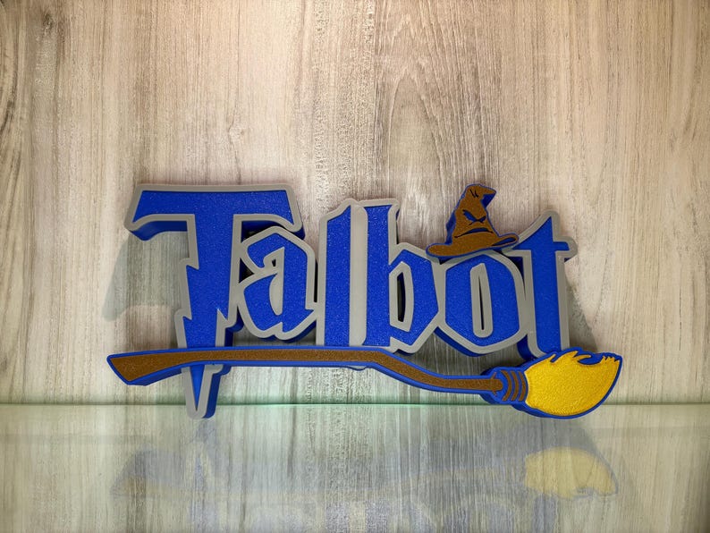 May include: A blue and silver 3D sign with the name "Talbot" in a stylised font. A brown and yellow broom with a wizard's hat is attached to the sign.