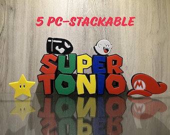 May include: A 5-piece stackable set featuring the text SUPER TONIO in red, green, blue, and yellow block letters. The set includes a black bullet character, a white ghost character, a yellow star, and a red hat with a white M logo.