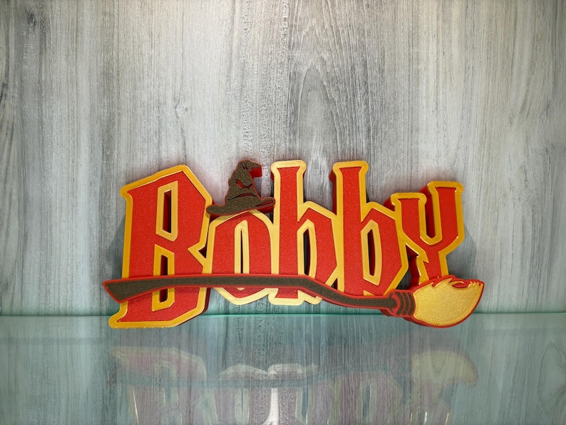 May include: Red and gold name sign with the name "Bobby" in a stylised font. A brown wizard's hat is above the "B" and a brown broomstick with a gold tip is below the name.