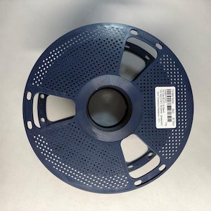 May include: A dark blue circular 3D printer filament spool with a perforated design and a central hub. A white rectangular label is affixed to the side, displaying technical specifications including 1kg, 1.75mm, and temperature settings.