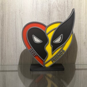 Può includere: Una targa di legno a forma di cuore con un design nero e grigio di due maschere di supereroi. Il lato sinistro è rosso e dice "BEST" e il lato destro è giallo e dice "FRIENDS".