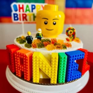 Puede incluir: Un pastel blanco decorado con gomitas de colores y una figura de Lego. El pastel está cubierto con un nombre con temática de Lego escrito en ladrillos de Lego de colores. El nombre es "JAINE".