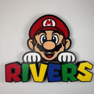 May include: A wall-mounted sign featuring a graphic of Mario from the Super Mario franchise with white gloved hands resting on top of the word RIVERS. The letters are rendered in a blocky, multi-coloured style with red, green, blue, and yellow.