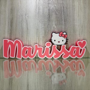 Puede incluir: Un letrero de nombre 3D rosa con el nombre "Marissa" y un personaje de Hello Kitty con un lazo rojo en la parte superior. El letrero está sobre un fondo de madera.