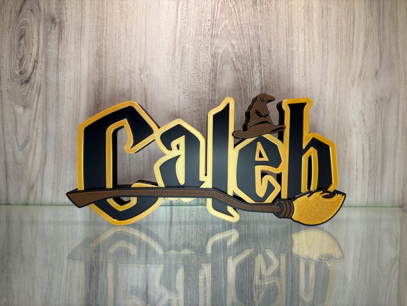 May include: A black and gold wooden sign with the name "Caleb" in a stylised font. A brown wizard's hat sits above the "a" in Caleb. A brown and gold broomstick is attached to the bottom of the sign.
