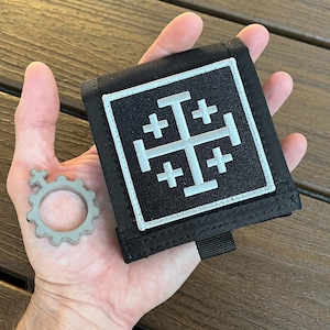 May include: A black square fabric patch featuring an embroidered white Jerusalem cross design with four smaller crosses in the corners, accompanied by a separate circular metal gear-shaped charm with a small cross symbol at the top edge.