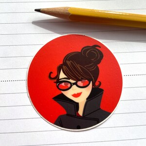 May include: A red circle sticker with a cartoon illustration of a woman with brown hair, wearing red sunglasses and a black coat. The woman is smiling.