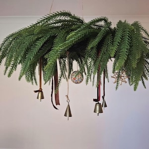 May include: A hanging circular wreath made of dense green pine needles. Suspended from the foliage are several metallic bells, a small round ornament with a floral pattern, a straw star, and thin brown ribbons hanging at varying lengths.