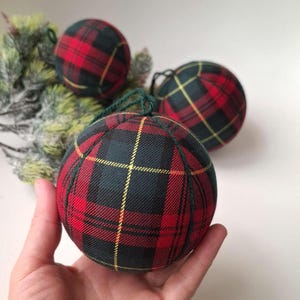 May include: Three spherical ornaments covered in a red, black, and yellow tartan fabric pattern. Each ornament features dark green fabric seams and a matching dark green string loop at the top for hanging. The fabric has a visible woven texture.