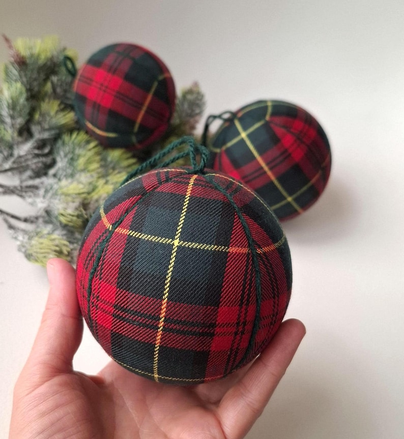 May include: Three spherical ornaments covered in a red, black, and yellow tartan fabric pattern. Each ornament features dark green fabric seams and a matching dark green string loop at the top for hanging. The fabric has a visible woven texture.