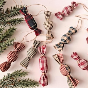 Puede incluir: Una colección de adornos de tela con forma de caramelos envueltos, con varios patrones de cuadros y tartán en rojo, negro, beige y verde. Cada adorno incluye un lazo de cuerda de yute para colgar, acompañado de ramas de pino verdes.