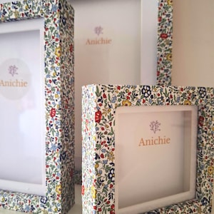Può includere: Tre cornici per foto con bordi in tessuto floreale. Le cornici sono bianche con un motivo floreale colorato in rosso, blu, giallo e verde. Il marchio "Anichie" è stampato sullo sfondo bianco delle cornici.