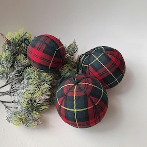 May include: Three spherical ornaments covered in a red, dark green, and yellow plaid fabric pattern. Each ornament features a dark green string loop at the top for hanging. The fabric is segmented by dark green stitching lines across the surface.