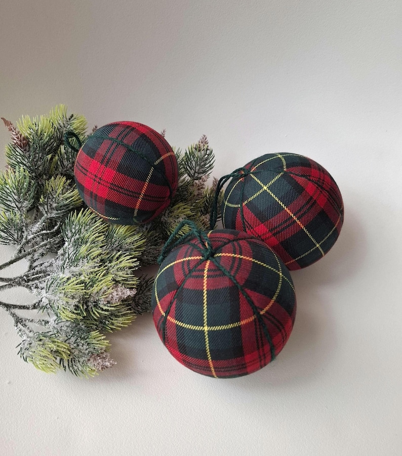 May include: Three spherical ornaments covered in a red, dark green, and yellow plaid fabric pattern. Each ornament features a dark green string loop at the top for hanging. The fabric is segmented by dark green stitching lines across the surface.