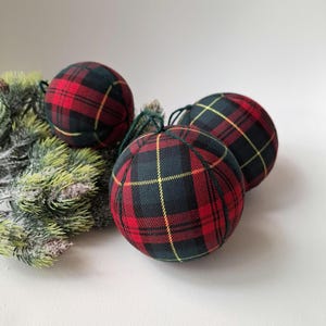May include: Three spherical baubles covered in a red, black and yellow tartan fabric pattern. Each bauble features a dark green string loop at the top for hanging. The baubles are arranged near a cluster of artificial frosted pine needles.