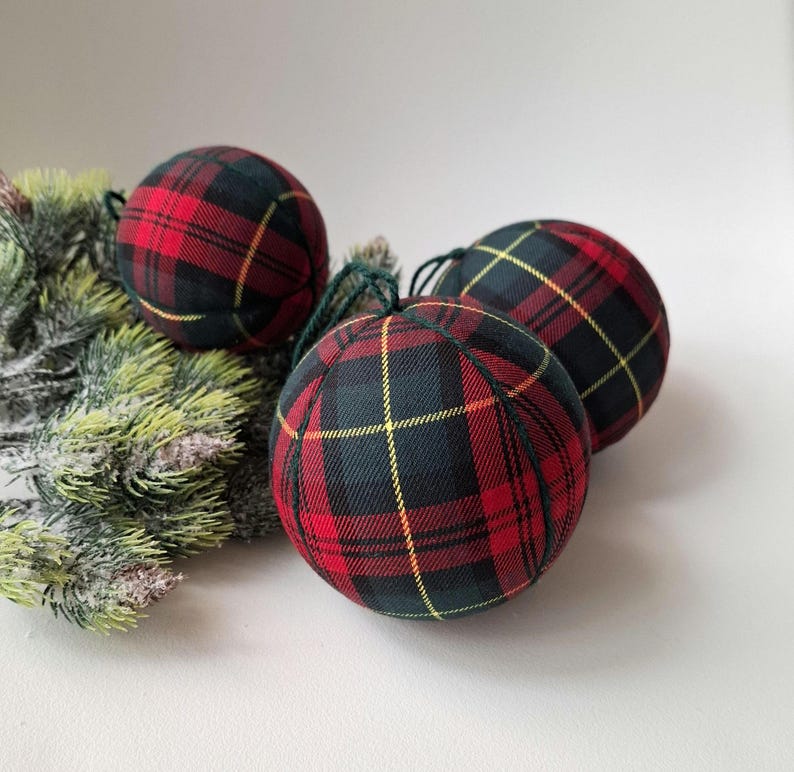 May include: Three spherical baubles covered in a red, black and yellow tartan fabric pattern. Each bauble features a dark green string loop at the top for hanging. The baubles are arranged near a cluster of artificial frosted pine needles.