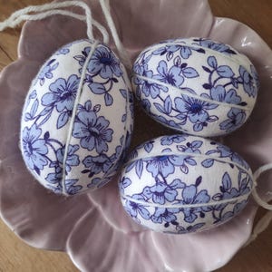 May include: Three white fabric Easter eggs with blue floral patterns. The eggs are sitting on a light purple plate.