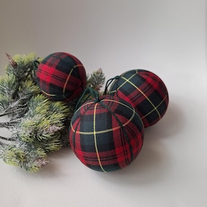 May include: Three spherical baubles covered in a red, black and yellow tartan fabric pattern. Each bauble features a dark green string loop at the top for hanging. The baubles are arranged near a small cluster of artificial pine branches.