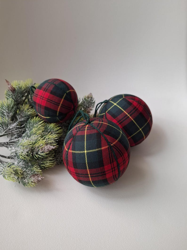 May include: Three spherical baubles covered in a red, black and yellow tartan fabric pattern. Each bauble features a dark green string loop at the top for hanging. The baubles are arranged near a small cluster of artificial pine branches.