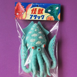 May include: A teal blue felt squid plush toy with red eyes and white clouds on its body. The toy is packaged in a clear plastic bag with a colourful label that reads "怪獣 アタック" in Japanese.