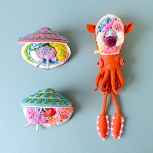 Op de afbeelding: Drie vilt sculpturen van zeedieren. Een rode en oranje inktvis met een gedetailleerd interieur, en twee schelpen met kleurrijke geborduurde interieurs.