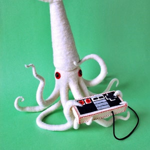 K&ouml;nnte beinhalten: Ein wei&szlig;er Filz-Tintenfisch mit roten Augen h&auml;lt einen schwarzen und grauen Videospiel-Controller. Der Tintenfisch hat einen langen, spitzen Kopf und acht Tentakel.