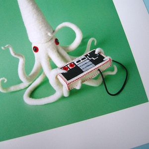 K&ouml;nnte beinhalten: Ein wei&szlig;er Filz-Oktopus mit roten Knopfaugen, der einen alten Videospiel-Controller mit einem schwarzen Kabel spielt.