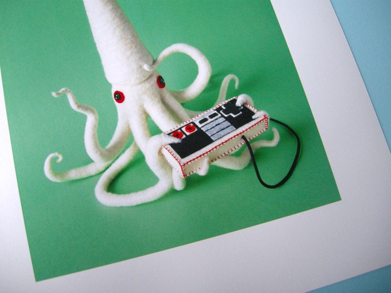 K&ouml;nnte beinhalten: Ein wei&szlig;er Filz-Oktopus mit roten Knopfaugen, der einen alten Videospiel-Controller mit einem schwarzen Kabel spielt.