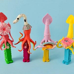 Puede incluir: Cuatro figuritas de fieltro de pulpo y calamar con cuerpos y sombreros de diferentes colores. Cada figurita está de pie sobre una base texturizada y colorida.