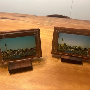 May include: Two wooden picture frames with a cityscape photo inside. The frames are brown and have a clear acrylic front. The frames are standing on a wooden table.