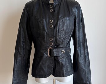 May include: A black leather jacket featuring a high collar, long sleeves, and two chest pockets. The front closure includes a series of metal eyelets and a matching black leather belt with a rectangular metal buckle at the waist area.