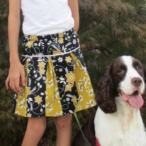 May include: A young girl wearing a white shirt and a black and yellow pleated skirt with floral print. The skirt has a white trim. The girl is standing next to a brown and white dog. The text "aivilo pleated skirt" and "Sizes 6-12m to 14 tween" is visible in the image.