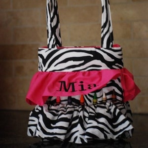 May include: A black and white zebra print tote bag with a pink ruffle trim and the name "Mia" embroidered on the front. The bag has a zipper closure and is filled with colorful crayons.