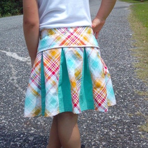 Puede incluir: Una niña pequeña con una camiseta blanca y una falda de cuadros con rayas de color turquesa, rojo, amarillo y blanco. La falda tiene una cintura blanca y está plisada.