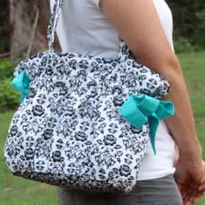 Puede incluir: Una mujer lleva un bolso tote floral blanco y negro con lazos turquesa. El bolso tiene una correa larga y se lleva sobre el hombro.