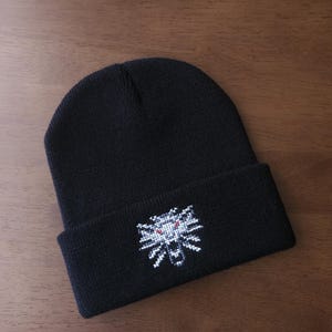 May include: Black beanie hat featuring a white and red embroidered wolf emblem. The hat is made of a soft, knitted material, ideal for colder weather. The hat is displayed on a wooden surface.