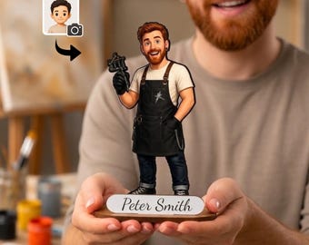 May include: A custom cartoon-style figurine of a person with a beard, wearing a white t-shirt, black apron, and dark jeans, holding a tattoo machine. The figure stands on a wooden base with a white nameplate that reads Peter Smith.